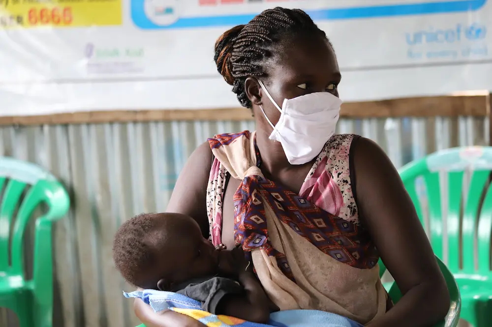 Nyaluit Khan and her 14-month-old child Chudier at a Concern Nutrition Center in South Sudan to receive supplementary food such as Plumpynut and cereal to help increase their nutritional levels. Nyaluit wears a mask while in the waiting areas of the centre to help prevent the spread of COVID-19